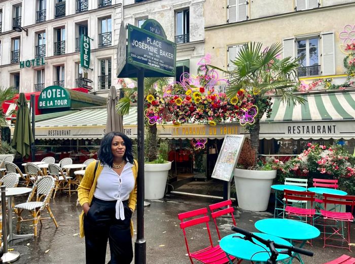 The author leans against a sign for Place Josephine Baker in Montparnasse, Paris, surrounded by cafe awnings and vibrant tables and chairs lining the sidewalk.