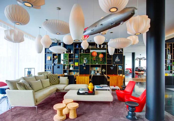 The lobby/lounge area of the citizenM Paris Charles de Gaulle Airport hotel is illuminated by numerous white hanging ceiling lights, featuring a sofa and two vibrant red chairs.