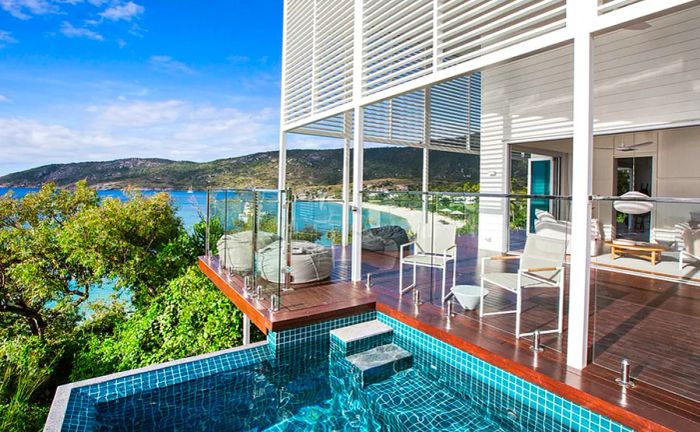 An outdoor deck furnished with white furniture overlooks a pool in the foreground and a beach in the background.