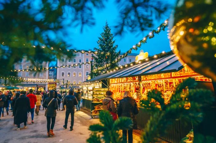 A Christmas market in Salzburg at sunset, with lights brightening the stalls and garlands of lights hanging overhead, while people in warm coats stroll about.