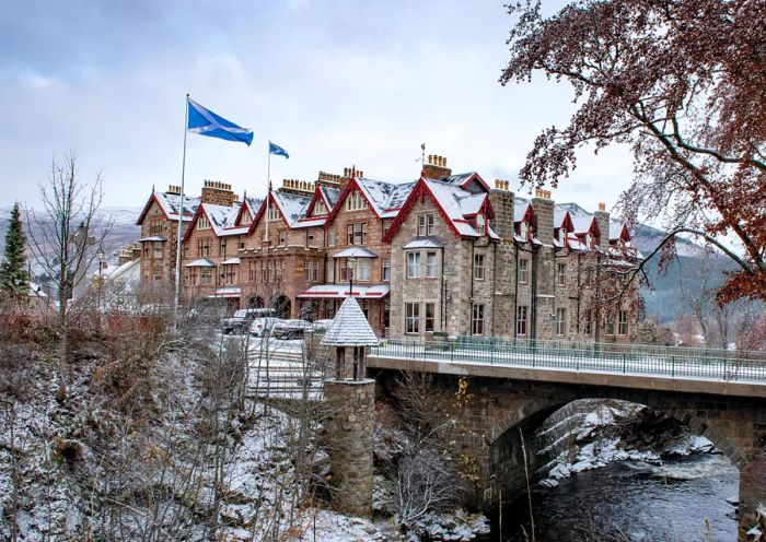 The Fife Arms hotel, blanketed in snow, perched by the River Cluny, adorned with two blue and white flags