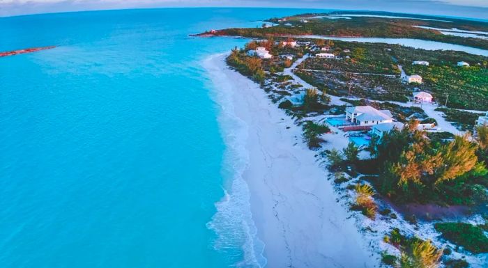 Aerial view of Exuma Island showcasing a long stretch of untouched beach, crystal-clear waters, patches of greenery, and a few houses with swimming pools.