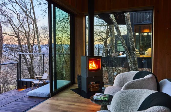 Imagine a tree house with an open sliding door revealing the stark beauty of bare trees outside; inside, two cozy chairs are positioned near a wood-burning stove.