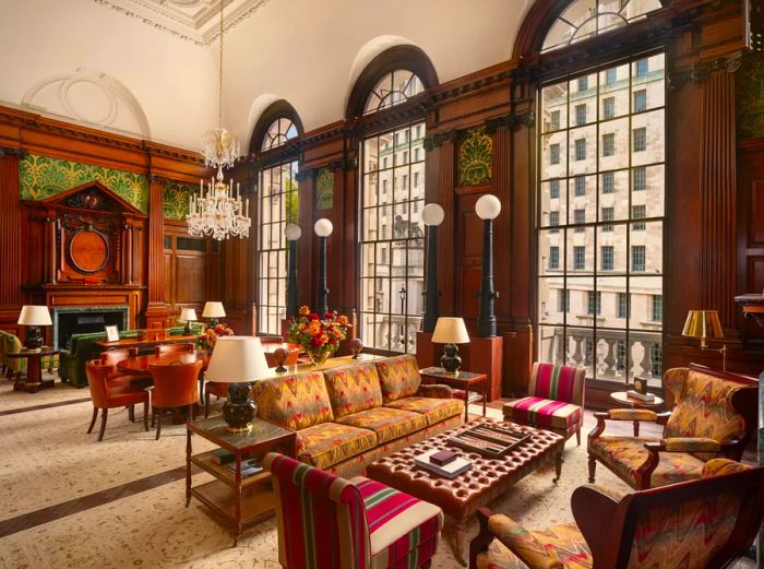 The Churchill Suite at Raffles London at the OWO features a spacious lounge area adorned with intricately carved wood accents, plush tufted leather furniture, and an elegant crystal chandelier.