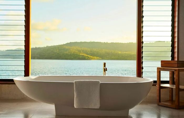 A freestanding white bathtub positioned next to a large window offers views of the water and green hills in the distance.