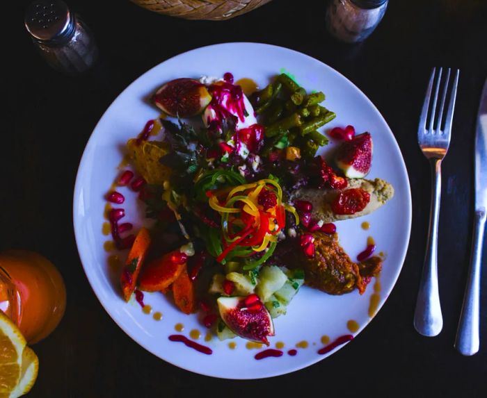 An overhead view of a white plate adorned with vibrant vegetables, figs, and pomegranate seeds, accompanied by a fork and knife.