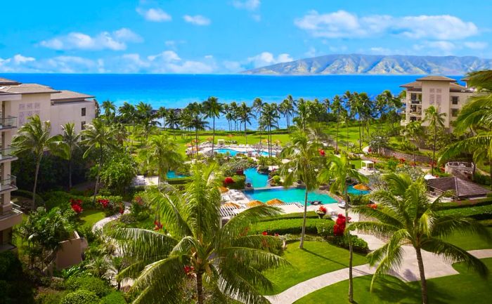 Aerial view of Montage Kapalua Bay, showcasing lush lawns, palm trees, multiple pools, and beige buildings adjacent to the ocean.