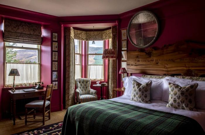 A guest room at the Fife Arms features a wooden desk, an armchair nestled in a curtained window nook, a Scottish tartan throw draped over the bed, and charming wood floors.