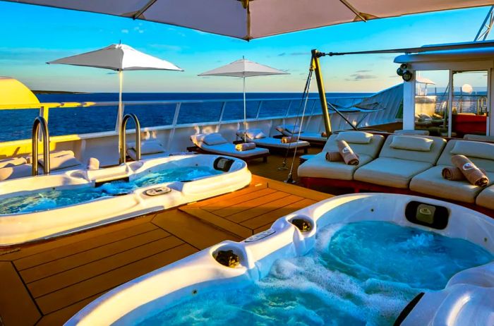 Two hot tubs on the upper deck of a SeaDream Yacht Club vessel, surrounded by lounge chairs and umbrellas.