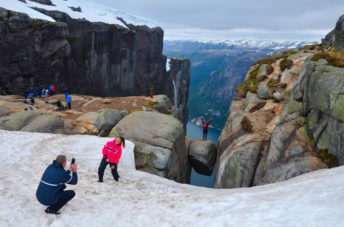 A woman in a pink jacket smiles for a photo near a cliff, while another woman stands behind her, arms outstretched on a suspended rock.