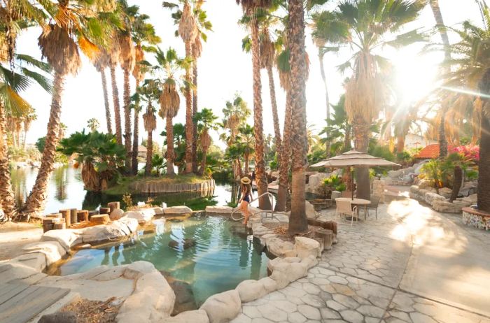 A woman walks into one of the 50 geothermal pools at Murrieta Hot Springs, surrounded by palm trees, stones, and winding pathways.