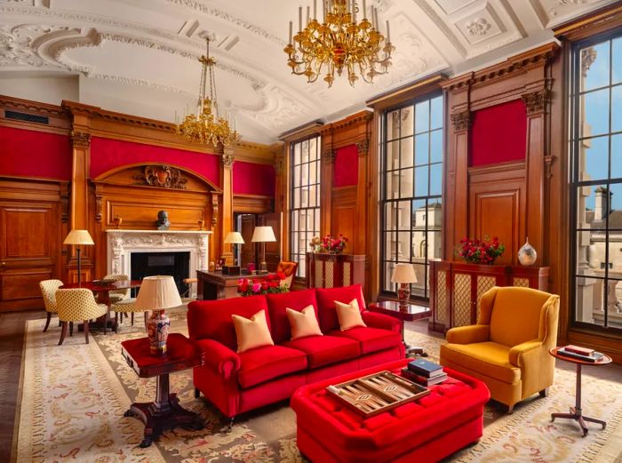 The Haldane Suite at Raffles London at The Owo boasts dark wood paneling, a striking red sofa, and ornate gilt chandeliers.