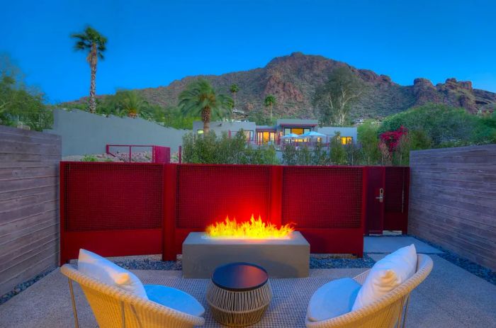 Two chairs positioned in front of a glowing outdoor firepit, framed by a red wall, with palm trees and a mountain visible in the background.