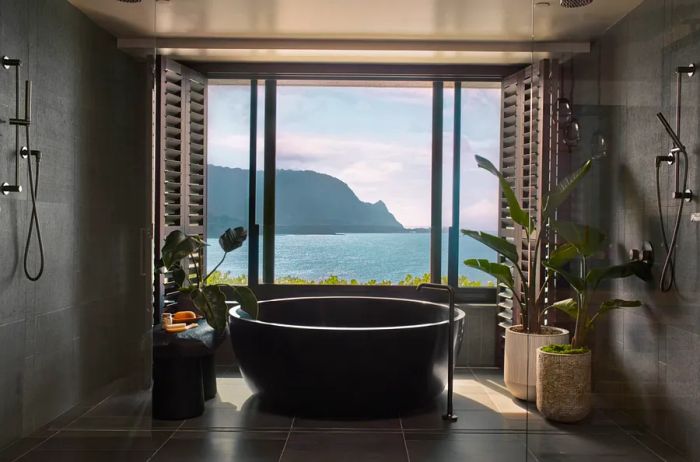 The bathroom in the Napali Penthouse suite features lush plants and a spacious round tub positioned next to a large window offering ocean views.