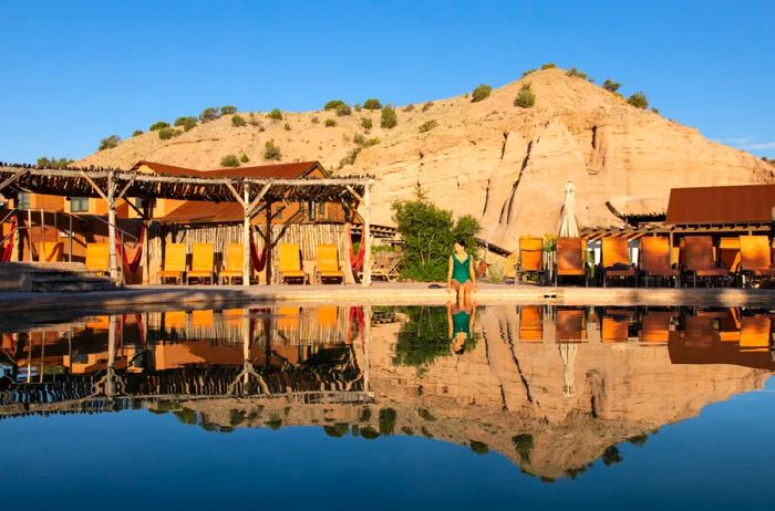 A woman relaxes at the edge of a mineral pool at Ojo Caliente, framed by a rocky mountain backdrop and lounge chairs.