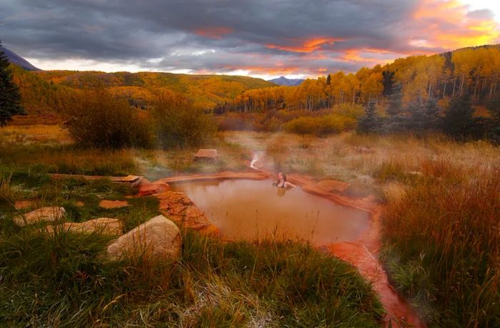 An individual enjoying a soak in an outdoor mineral pool at Dunton Hot Springs, surrounded by trees adorned in autumn colors.