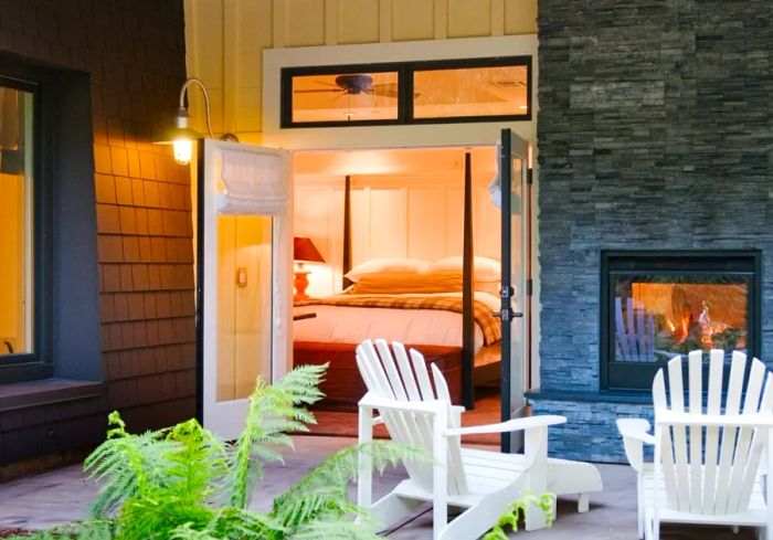 A view of a guest room at the Farmhouse Inn from the terrace, showcasing open doors, a cozy fireplace, and two white Adirondack chairs.