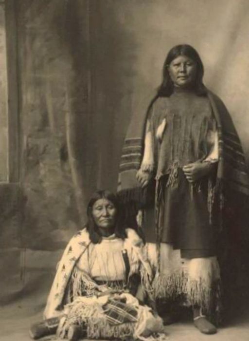 A black and white photo featuring two Native American women, one sitting and the other standing.