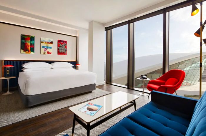 A guest room at the TWA Hotel featuring blue and red decor, hardwood floors, and expansive floor-to-ceiling windows.