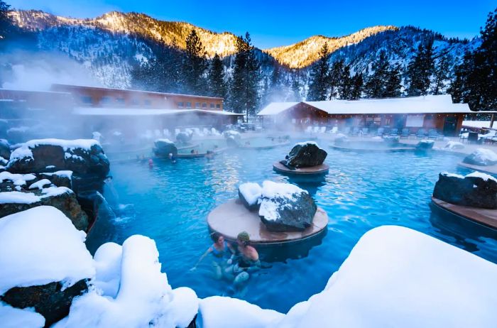 Guests enjoying the steaming hot springs at Quinn's Hot Springs Resort, with snow blanketing the ground and rooftops, and majestic mountains in the background.