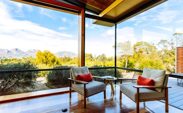 A cozy corner of a covered wooden deck features two vacant white armchairs with a picturesque countryside backdrop.