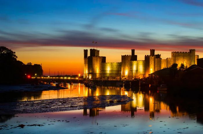 A medieval castle at sunset, dramatically illuminated against a backdrop of a pink, gold, and blue sky, all mirrored in the water below.