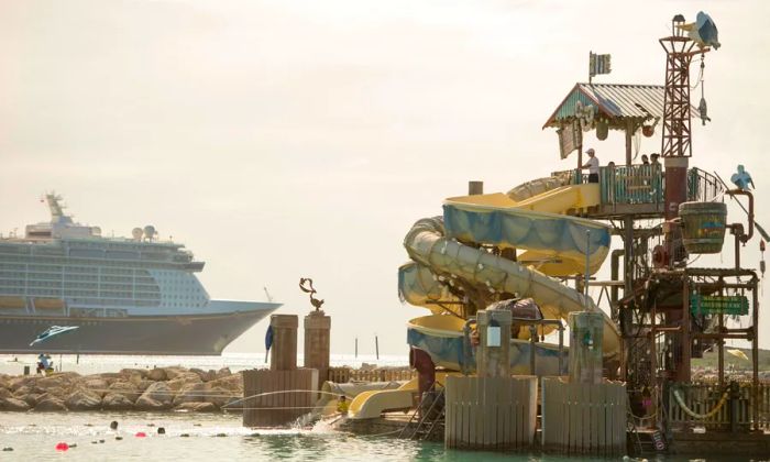 A pelican-themed waterslide in the foreground on Castaway Cay, with a Disney Cruise Line ship visible in the background.