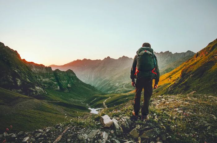 The back view of a person wearing a backpack as they trek through a mountainous valley.