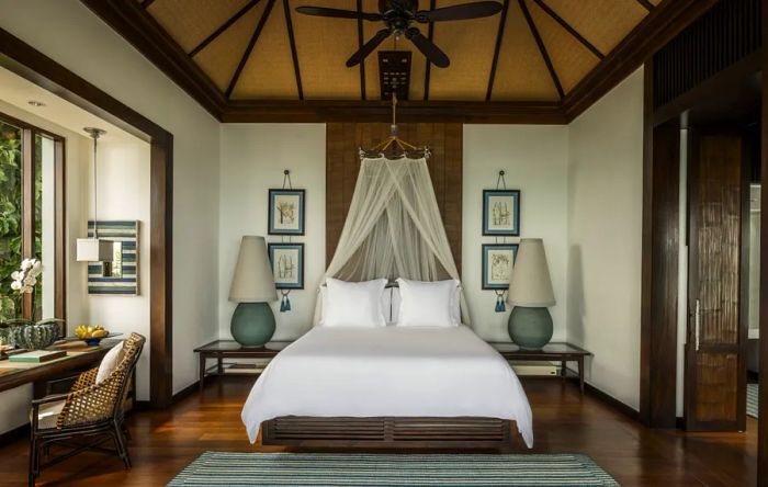 A bedroom at the Four Seasons Resort Koh Samui features white walls, dark wood flooring, netting tucked behind the bed, and soaring ceilings.