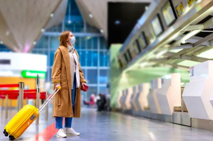 A person wearing a long brown coat and a mask, holding a yellow rolling suitcase, gazes at the departures board in an airport.