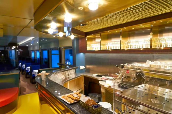 An empty dining car on a Treni Turistici Espresso train features a few cookies on the counter, glasses arranged on shelves behind the service area, and vacant booths.