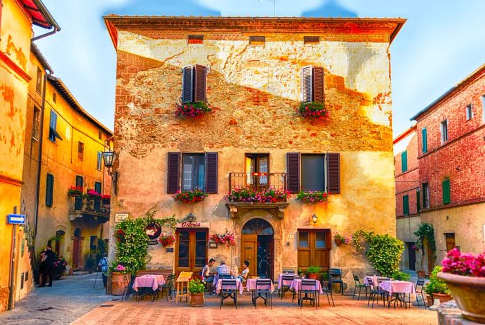 An ancient structure in the medieval Tuscan village of Pienza features a restaurant with an outdoor terrace, furnished with tables and chairs, where several patrons enjoy their meals on the ground floor.