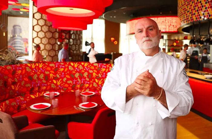 A man dressed in a white chef's uniform with a white beard stands before a red circular booth, featuring an open kitchen and a restaurant in the background.