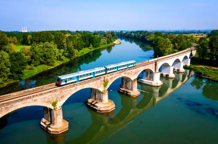 Aerial view of a vintage turquoise and white train crossing a river on a bridge adorned with multiple arches