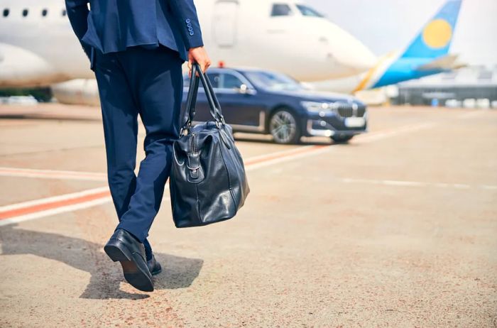 A person dressed in a blue suit carries a leather weekender as they walk across the tarmac toward a plane.