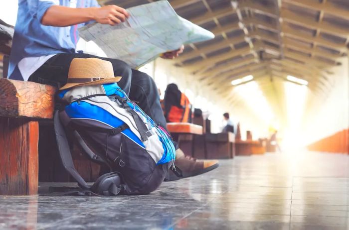 A traveler examines a paper map while seated on a bench in what appears to be a train station, their backpack resting beside them.