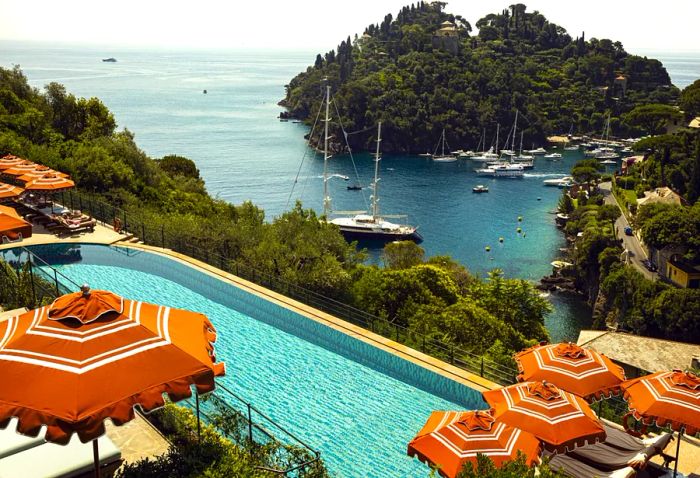 A cliffside swimming pool adorned with orange umbrellas, offering views of a harbor filled with sailboats.