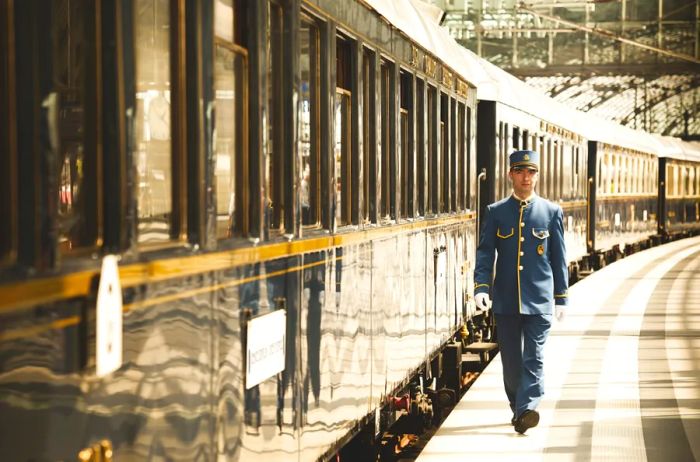 A man dressed in a blue uniform with gold accents stands next to a sleek line of gleaming train cars inside a train station.