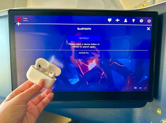 A woman holding a pair of AirPods Pro in front of an airplane TV screen displaying 'Bluetooth,' followed by 'refresh' and 'please select a device below or refresh to search again.'