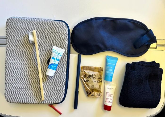 An overhead view of Air France's amenity kit showcases a compostable toothbrush, toothpaste, a pen, socks, an eye mask, ear plugs, and two moisturizers from Clarins, all neatly arranged on a small toiletry bag with a blue and white herringbone pattern.