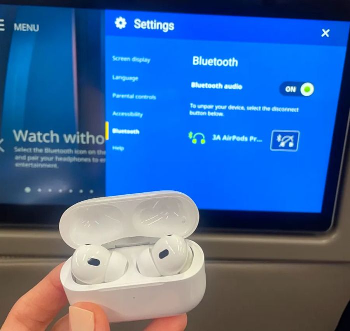 A hand holding AirPods Pro in their case next to the Bluetooth technology screen onboard United flights.