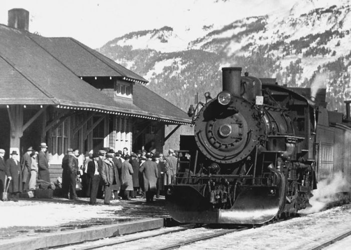 A historic photo of the Alaska Railroad engine at the station platform, featuring passengers.