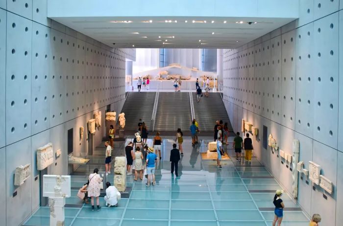 An interior view of the Acropolis Museum in Athens, featuring visitors exploring ancient sculptures and artifacts.