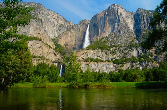 A dramatic rocky cliff with two waterfalls cascading into a lush green riverbed: Upper and Lower Yosemite Falls.