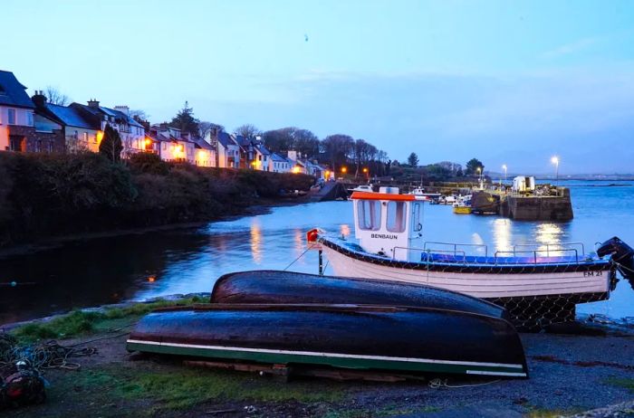 Three small boats resting on the shore, with distant houses silhouetted against the dusk sky.