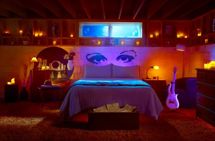 Prince's Purple Rain house showcasing a purple-lit bed accompanied by a guitar and stand nearby, with artistic eyes drawn on the wall behind and candles surrounding the space.