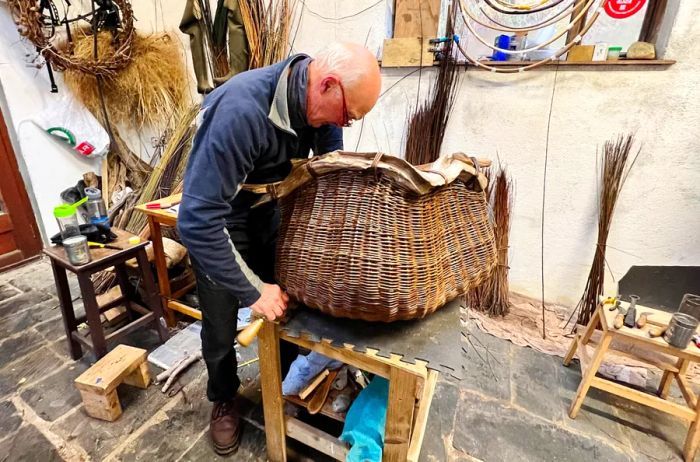 A basket maker hunched over a large brown basket in his workshop