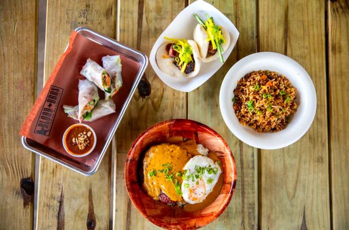 An overhead shot of spring rolls, brisket fried rice, loco moco, and brisket-stuffed bao at Blood Bros. BBQ, displayed on a wooden table.