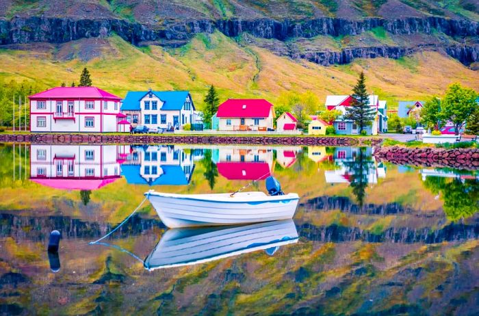 A small fishing boat glides on the water, framed by the vibrant village of Seydisfjordur, Iceland, against a backdrop of rugged cliffs and lush greenery.