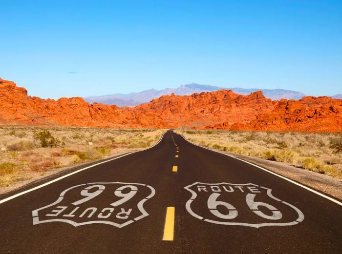 Route 66 pavement sign against a backdrop of Mojave Desert red rock mountains.